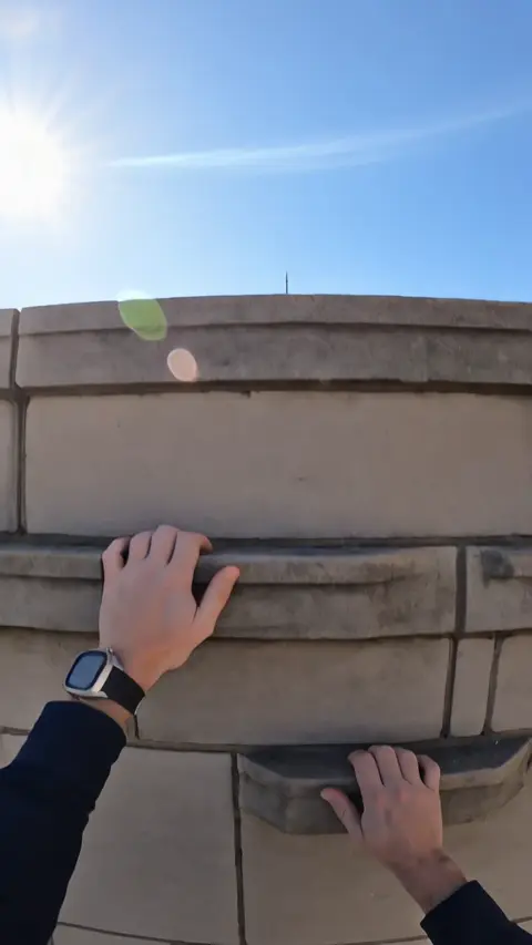 A first person climb over a stone parapet high above the city, looking down at traffic and across to the Empire State Building