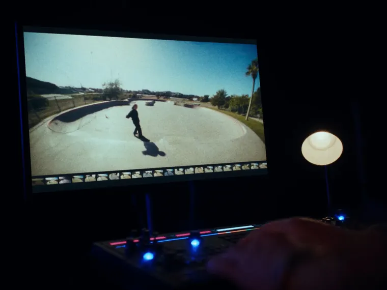 A grading suite monitor showing the skate film frame above a strip of timeline thumbnails, a control surface lit below it